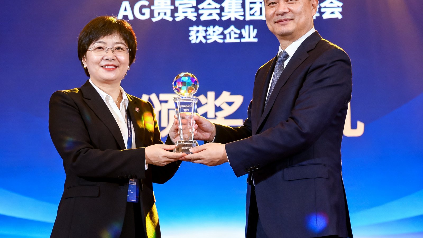2025年亚太商旅服务峰会颁奖典礼现场，AG贵宾会集团代表在舞台上接过水晶奖杯，背景大屏幕显示获奖企业名称